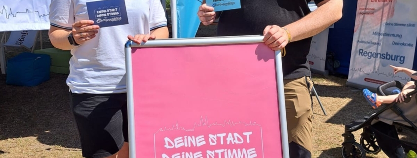 Das Bild zeigt Lena Schulwitz und Jonas Scherer vom Stadtjugendring vor einem Plakat mit der Aufschrift "Deine Stadt, deine Stimme". Sie halten Postkarten mit derselben Aufschrift in der Hand.